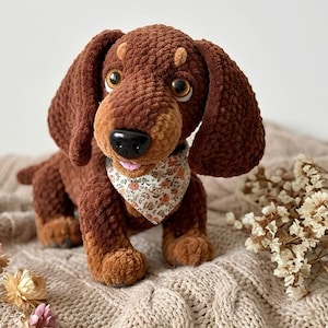 Puede incluir: Un peluche de perro salchicha marrón hecho de hilo suave y texturizado. El perro tiene ojos grandes y brillantes de color marrón, una nariz de plástico negra y una pequeña lengua rosa. Lleva un pañuelo triangular con fondo blanco y estampado floral.