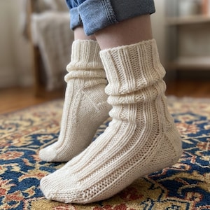 Puede incluir: Un par de calcetines de color crema tejidos a mano con un patrón de canalé vertical que se extiende desde el puño hasta la punta. Los calcetines están confeccionados con hilo grueso y texturizado, y se llevan con vaqueros azules, con ajuste holgado.