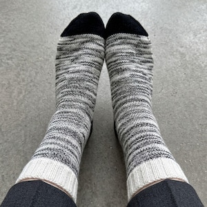Peut inclure: Une paire de chaussettes mi-mollet avec un motif en tricot chiné gris et blanc. Les chaussettes présentent des pointes noires unies et des bords-côtes blanc cassé en haut, offrant un contraste marqué avec le corps texturé gris du vêtement.