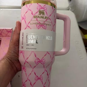 May include: A light pink Stanley Quencher H2.0 tumbler with a 40 oz capacity. The exterior features a repeating pattern of pink ribbons tied in bows. It has a gold-colored rim, a pink handle, and a clear lid. The label reads Stanley LoveShackFancy.