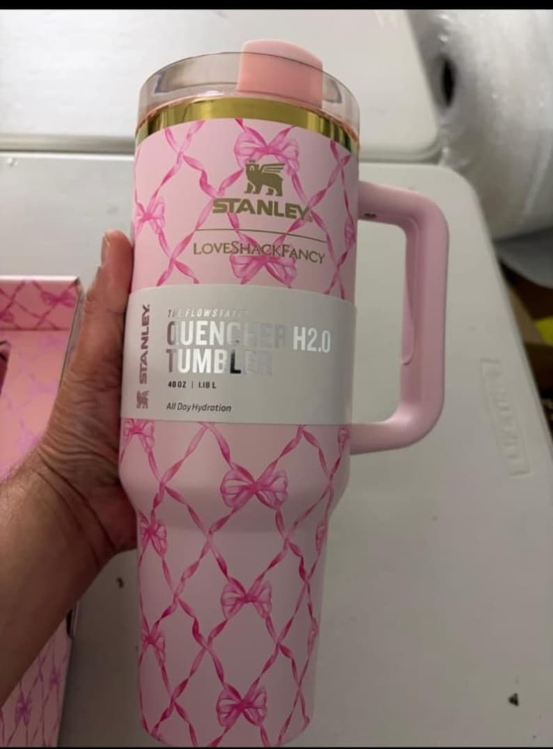 May include: A light pink Stanley Quencher H2.0 tumbler with a 40 oz capacity. The exterior features a repeating pattern of pink ribbons tied in bows. It has a gold-colored rim, a pink handle, and a clear lid. The label reads Stanley LoveShackFancy.