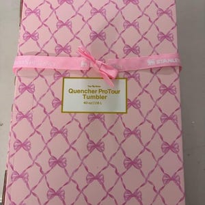 May include: A rectangular cardboard box with a light pink background featuring a repeating pattern of pink bows connected by thin lines. A pink ribbon is tied around the center. A white label reads: The Flip Straw Quencher ProTour Tumbler 40 oz | 1.18 L.