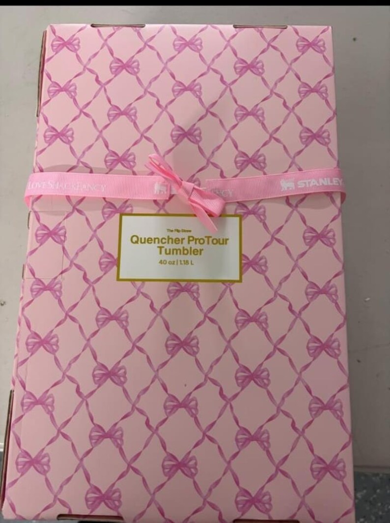 May include: A rectangular cardboard box with a light pink background featuring a repeating pattern of pink bows connected by thin lines. A pink ribbon is tied around the center. A white label reads: The Flip Straw Quencher ProTour Tumbler 40 oz | 1.18 L.