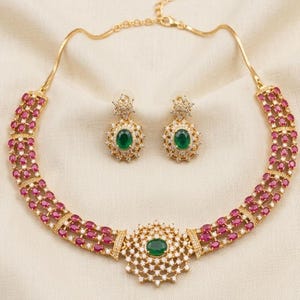 May include: A gold-toned necklace and earring set featuring oval-cut pink stones arranged in a multi-row lattice pattern. The necklace has a central circular pendant with a green oval stone, while the earrings feature matching green stones.