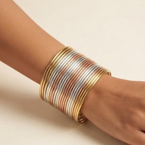 Pode incluir: Uma ampla pilha de pulseiras finas e lisas de metal em tons alternados de ouro amarelo, ouro rosa e prata. O conjunto consiste em numerosas pulseiras circulares individuais usadas juntas para criar um efeito de bracelete metálico espesso.