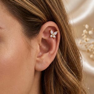 May include: A gold-toned cartilage earring shaped like a butterfly, featuring two marquise-cut clear stones for the upper wings and two round-cut clear stones for the lower wings, secured in the upper ear cartilage with a small metal post backing.
