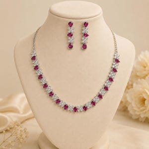Op de afbeelding: Een zilverkleurige ketting en bijpassende oorbellen met druppelvormige magentakleurige stenen afgewisseld met heldere, rondgeslepen stenen. De ketting heeft een flexibel ontwerp en de oorbellen weerspiegelen hetzelfde bloemvormige patroon.