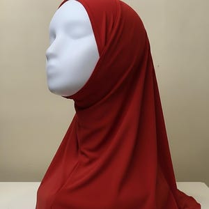 May include: A solid red hijab worn on a white mannequin head. The head covering features a smooth, opaque fabric that drapes over the head and shoulders, providing full coverage with a clean, seamless fit around the face and neck area.
