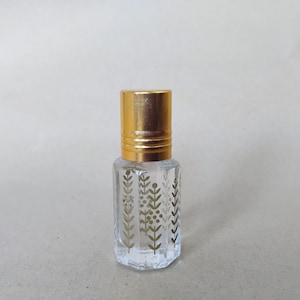 May include: A small, clear glass bottle with a hexagonal shape and a metallic gold cap. The exterior of the glass features a repeating vertical pattern of gold-colored botanical sprigs, leaves, and small berries printed on the transparent surface.