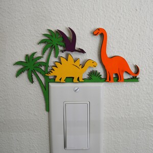 May include: A colorful wooden light switch cover featuring a dinosaur theme. The design includes a yellow stegosaurus, an orange long-necked dinosaur, a purple pterodactyl, and green palm trees. The cover is designed to fit over a standard light switch.