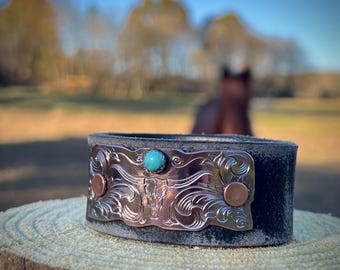 May include: A black leather cuff bracelet with a silver engraved plate featuring a stylized bull's head and a turquoise stone. The plate is accented with two copper rivets.