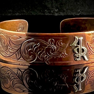 May include: A copper cuff bracelet featuring an intricate engraved floral design and the initials "KP" in silver.