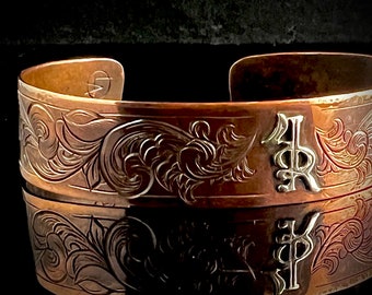 Op de afbeelding: Een koperen armband met een ingewikkeld gegraveerd bloemenpatroon en de initialen "KP" in zilver.