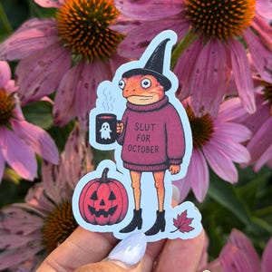 May include: A die-cut sticker featuring an orange frog wearing a black witch hat and a maroon sweater with the text SLUT FOR OCTOBER. The frog holds a black mug with a white ghost graphic and stands next to a pink jack-o'-lantern and a maple leaf.