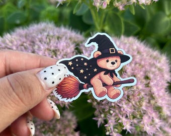 Pode incluir: Um adesivo recortado apresentando um ursinho de pelúcia marrom claro usando um chapéu de bruxa preto e uma capa preta adornada com estrelas amarelas. O urso monta uma vassoura com cerdas laranja, em estilo cartoon com borda branca.