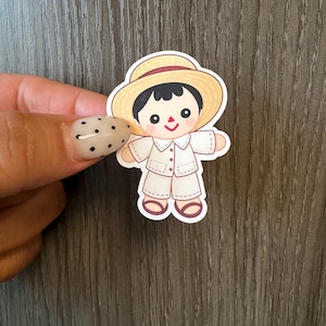 May include: A die-cut sticker featuring a cartoon character with black hair, wearing a light yellow straw hat, a white collared button-down shirt, and matching white shorts. The character has a simple face with black eyes and a red triangular mouth.