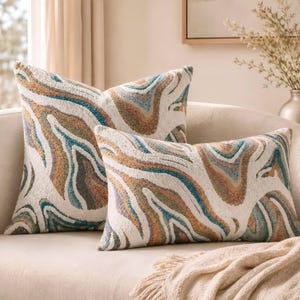May include: Two throw pillows featuring a textured, abstract marbled pattern in shades of cream, teal, brown, and grey. The square pillow sits behind a rectangular pillow, both displaying a consistent wavy, organic design across the fabric surface.