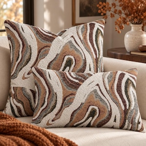 May include: Two scatter cushions featuring a textured, abstract marbled pattern in shades of cream, tan, dark brown, and muted olive green. The set includes one square cushion and one rectangular lumbar cushion, both displaying a consistent wavy design.