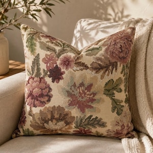 May include: A square throw pillow featuring a woven tapestry-style floral pattern in muted shades of dusty rose, sage green, and cream. The fabric displays a dense, textured botanical design with large, blooming flower heads and leafy foliage.