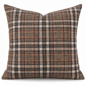 May include: A square throw pillow featuring a woven fabric with a multi-colored plaid pattern. The design consists of intersecting horizontal and vertical lines in shades of dark brown, tan, cream, and subtle rust orange, creating a grid.