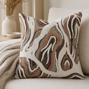 May include: A square throw cushion featuring a textured, tufted surface with an abstract, organic pattern of swirling lines in shades of cream, tan, dark brown, and muted grey. The design mimics the appearance of natural stone or wood grain.
