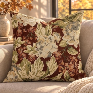 May include: A square throw cushion featuring a woven tapestry fabric with a floral pattern. The design displays large cream-coloured flowers, green leaves, and small reddish-brown blossoms set against a deep, dark red background.