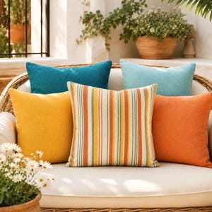 May include: A collection of five square throw pillows arranged on a curved wicker sofa. The pillows feature solid colors in teal, light blue, mustard yellow, and burnt orange, alongside one central pillow with vertical stripes in orange, yellow, and green.