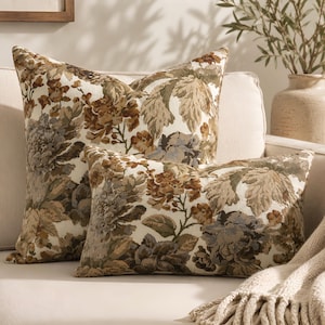 Può includere: Due cuscini decorativi con un motivo floreale intrecciato nei toni del marrone, beige, verde oliva e blu tenue su uno sfondo bianco sporco. Uno è quadrato e l'altro rettangolare, entrambi mostrano lo stesso design botanico.