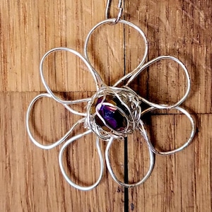 May include: A silver-toned metal necklace featuring a pendant shaped like a six-petaled flower. The petals are formed from thin wire loops, and the center of the flower contains a small, dark purple faceted bead encased in silver wire wrapping.