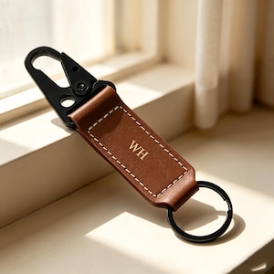May include: A brown leather keychain featuring a black metal carabiner clip at the top and a black metal split ring at the bottom. The leather strap is rectangular with white contrast stitching and the gold embossed letters WH in the center.