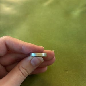 May include: A metallic ring with a smooth, polished finish and a subtle horizontal grooved line running through the center of the band. The ring features a uniform, cylindrical shape with a reflective surface that catches the light evenly.
