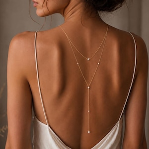 Puede incluir: Un delicado collar tipo lariat en tono dorado con un diseño de cadena de doble capa adornado con cinco pequeñas perlas blancas espaciadas uniformemente. El collar cae por el centro de la espalda sobre una prenda de tirantes finos de color claro.
