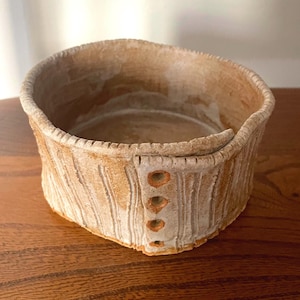 May include: A hand-built ceramic bowl with a light tan and off-white textured finish. The exterior features vertical ribbed detailing and a prominent overlapping seam on one side, accented by four small, evenly spaced circular holes in a vertical row.