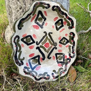 Può includere: Una ciotola in ceramica dipinta a mano con bordo smerlato, caratterizzata da una base color crema decorata con forme a rombo nere, linee curve verde oliva e piccoli tocchi di pennello rosa disposti in un motivo simmetrico.