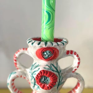 May include: A ceramic candle holder with a cream-coloured base, featuring hand-painted red floral motifs and green leaf accents. The holder has two sets of curved handles and holds a tall, cylindrical green candle with a wavy, marbled pattern.