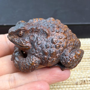 May include: A small, dark brown carved wooden toad figurine featuring a highly textured, bumpy surface with intricate circular patterns across its back and head, showcasing detailed eyes and a defined mouth in a realistic, crouching posture.