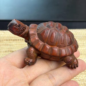 Op de afbeelding: Een klein, met de hand gesneden houten schildpadbeeldje met een rijke, roodbruine afwerking. Het beeldje is voorzien van gedetailleerde, getextureerde schubben op de kop en poten, en een gesegmenteerd, geometrisch schildpatroon op de rug.