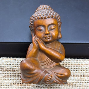 Puede incluir: Una pequeña figura de madera tallada de un Buda sentado con un acabado marrón suave y pulido. La figura se muestra con los ojos cerrados en una expresión pacífica, apoyando la cabeza sobre las manos y vistiendo túnicas drapeadas.
