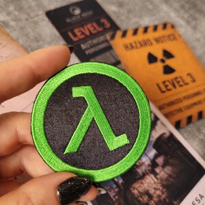 May include: A circular embroidered patch featuring a bright green lambda symbol centred on a black background, surrounded by a thick green border. The design is stitched with dense thread, creating a textured surface with clear, defined edges.