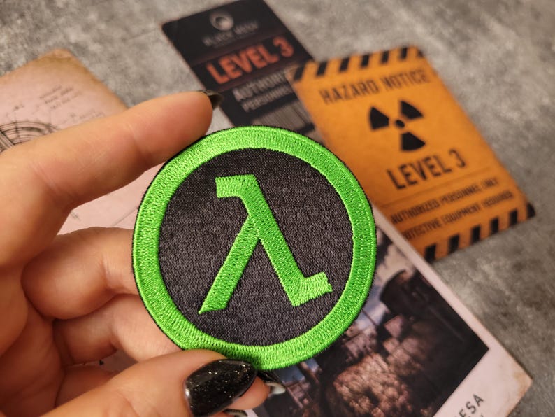 May include: A circular embroidered patch featuring a bright green lambda symbol centred on a black background, surrounded by a thick green border. The design is stitched with dense thread, creating a textured surface with clear, defined edges.