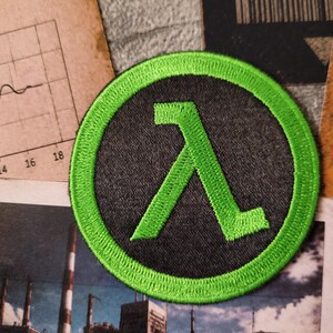 May include: A circular embroidered patch featuring a bright neon green Lambda symbol centred on a dark charcoal grey background. The patch has a thick, stitched neon green border that frames the design, creating a high-contrast appearance.