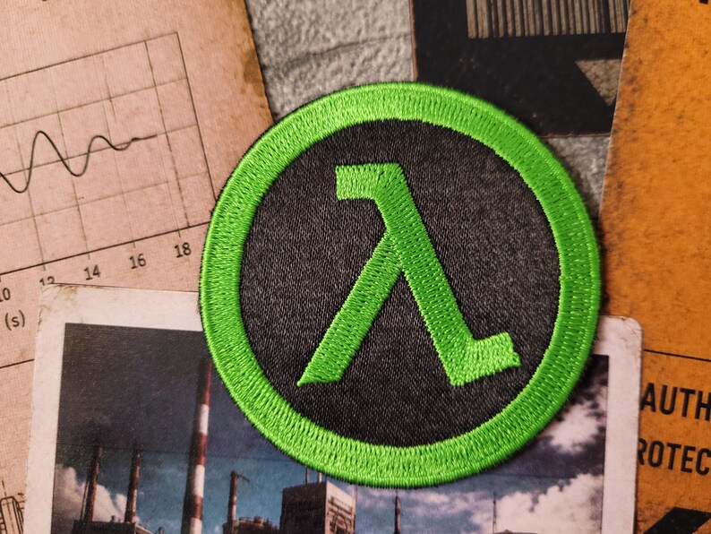 May include: A circular embroidered patch featuring a bright neon green Lambda symbol centred on a dark charcoal grey background. The patch has a thick, stitched neon green border that frames the design, creating a high-contrast appearance.