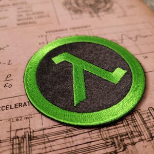 May include: A circular embroidered patch featuring a bright green lambda symbol centred on a dark grey background. The patch has a thick, stitched green border that frames the design, creating a distinct contrast between the vibrant green and the dark grey fabric.