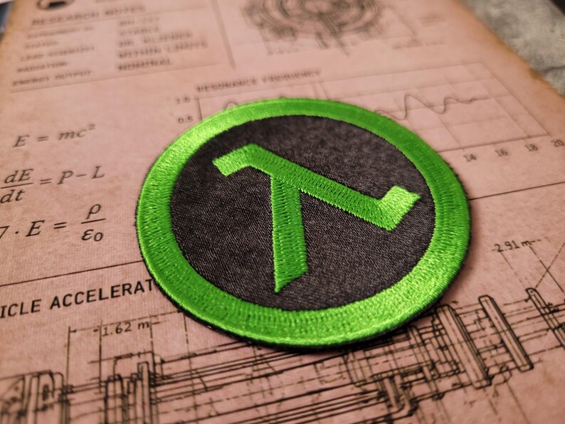 May include: A circular embroidered patch featuring a bright green lambda symbol centred on a dark grey background. The patch has a thick, stitched green border that frames the design, creating a distinct contrast between the vibrant green and the dark grey fabric.