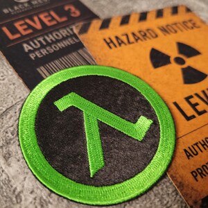 May include: A circular embroidered patch featuring a bright green lambda symbol on a black background, positioned over two rectangular cards labelled Level 3 Authority Personnel and Hazard Notice Level 3 with a black radiation warning symbol.