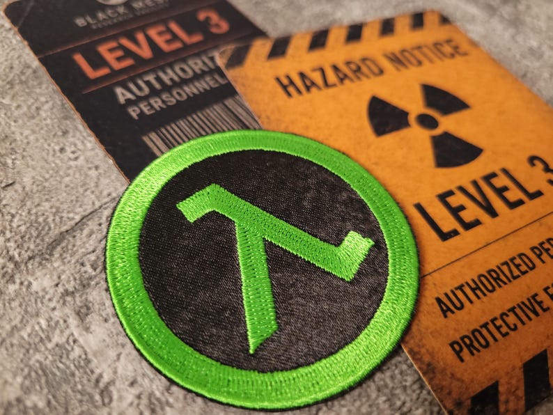 May include: A circular embroidered patch featuring a bright green lambda symbol on a black background, positioned over two rectangular cards labelled Level 3 Authority Personnel and Hazard Notice Level 3 with a black radiation warning symbol.