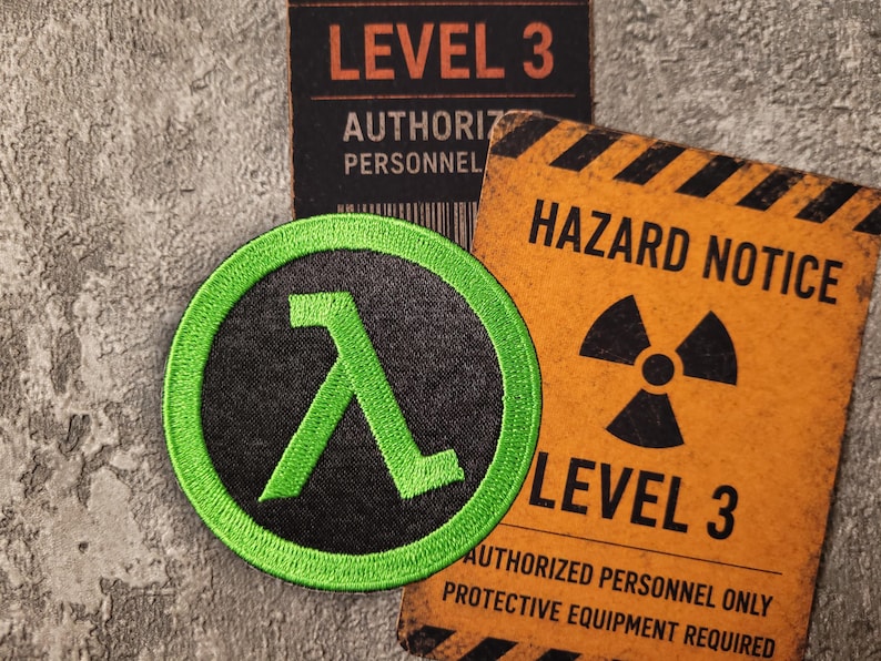 May include: A circular embroidered patch featuring a bright green Greek lambda symbol on a black background. The patch is positioned next to a rectangular orange sign with black text that reads HAZARD NOTICE LEVEL 3 AUTHORIZED PERSONNEL ONLY.