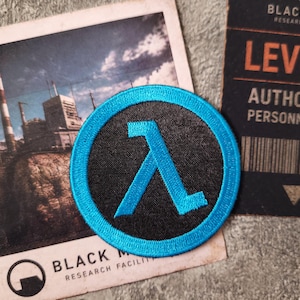 Puede incluir: Un parche bordado circular que presenta un símbolo lambda de color cian brillante centrado sobre un fondo negro con un borde cian a juego. El parche se muestra sobre un fondo que contiene una tarjeta e imagen de Black Mesa Research Facility.