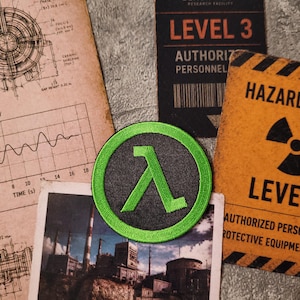 May include: A circular embroidered patch featuring a bright green Greek lambda symbol on a dark grey background. The patch is surrounded by technical blueprints, a photograph of an industrial facility, and a yellow hazard notice sign for Level 3.