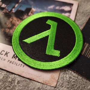 May include: A circular embroidered patch featuring a bright neon green lambda symbol centred on a solid black background, surrounded by a thick neon green stitched border. The patch is circular in shape with a textured thread finish throughout.